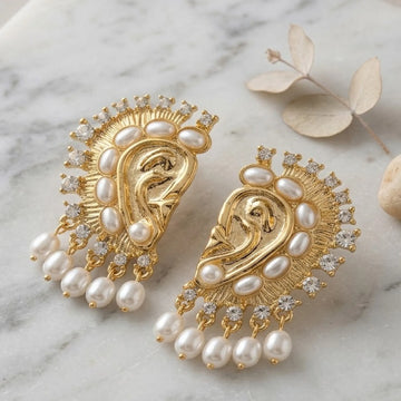 Gold and pearl earrings on a marble surface with dried leaves.