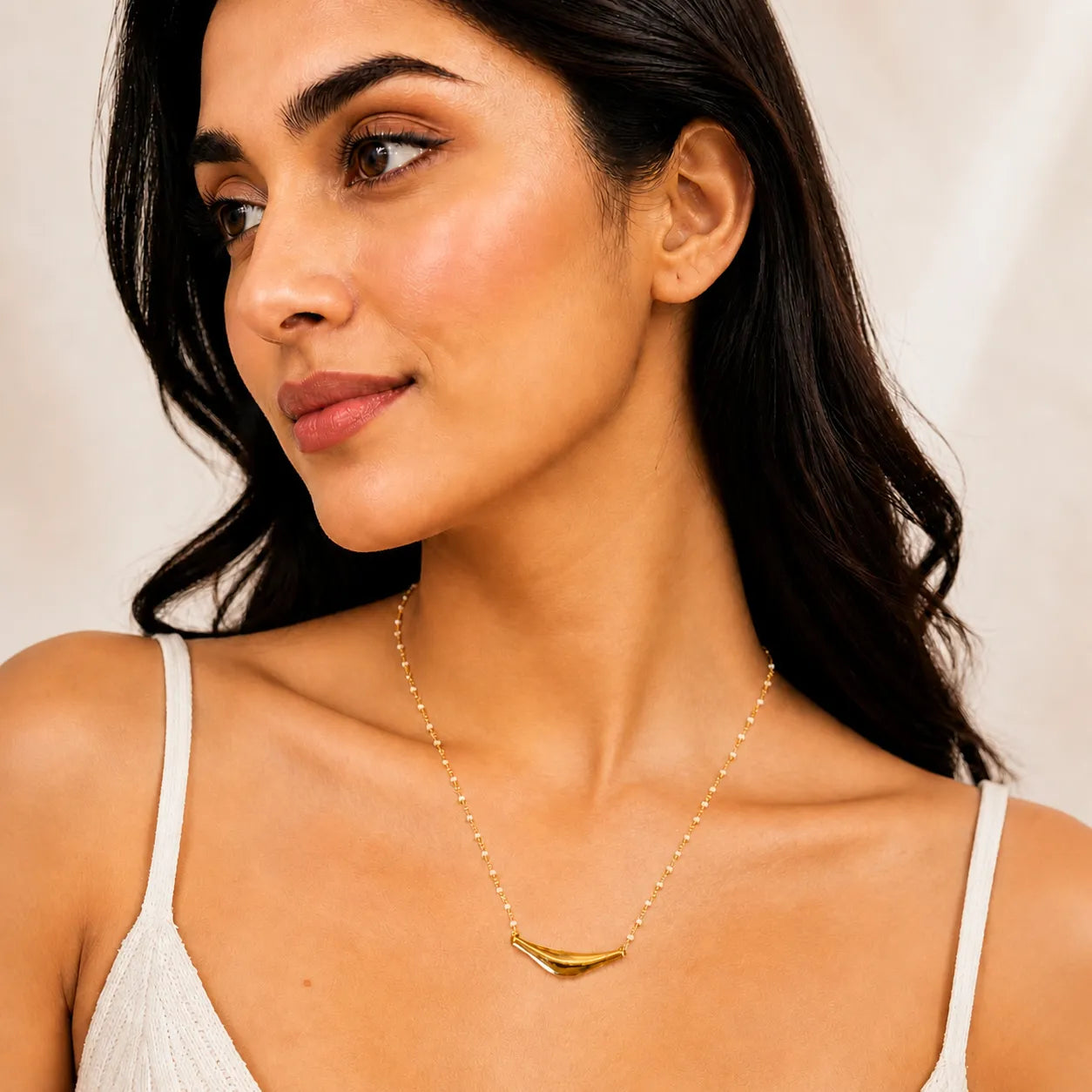 Woman wearing a gold necklace with a white top against a neutral background