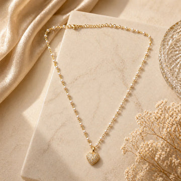 Gold necklace with a heart pendant on a marble surface with beige fabric and dried flowers.