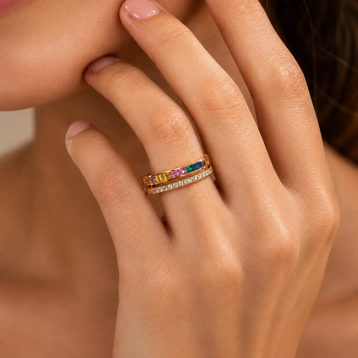 Close-up of a hand wearing a gold ring with colorful gemstones against a neutral background
