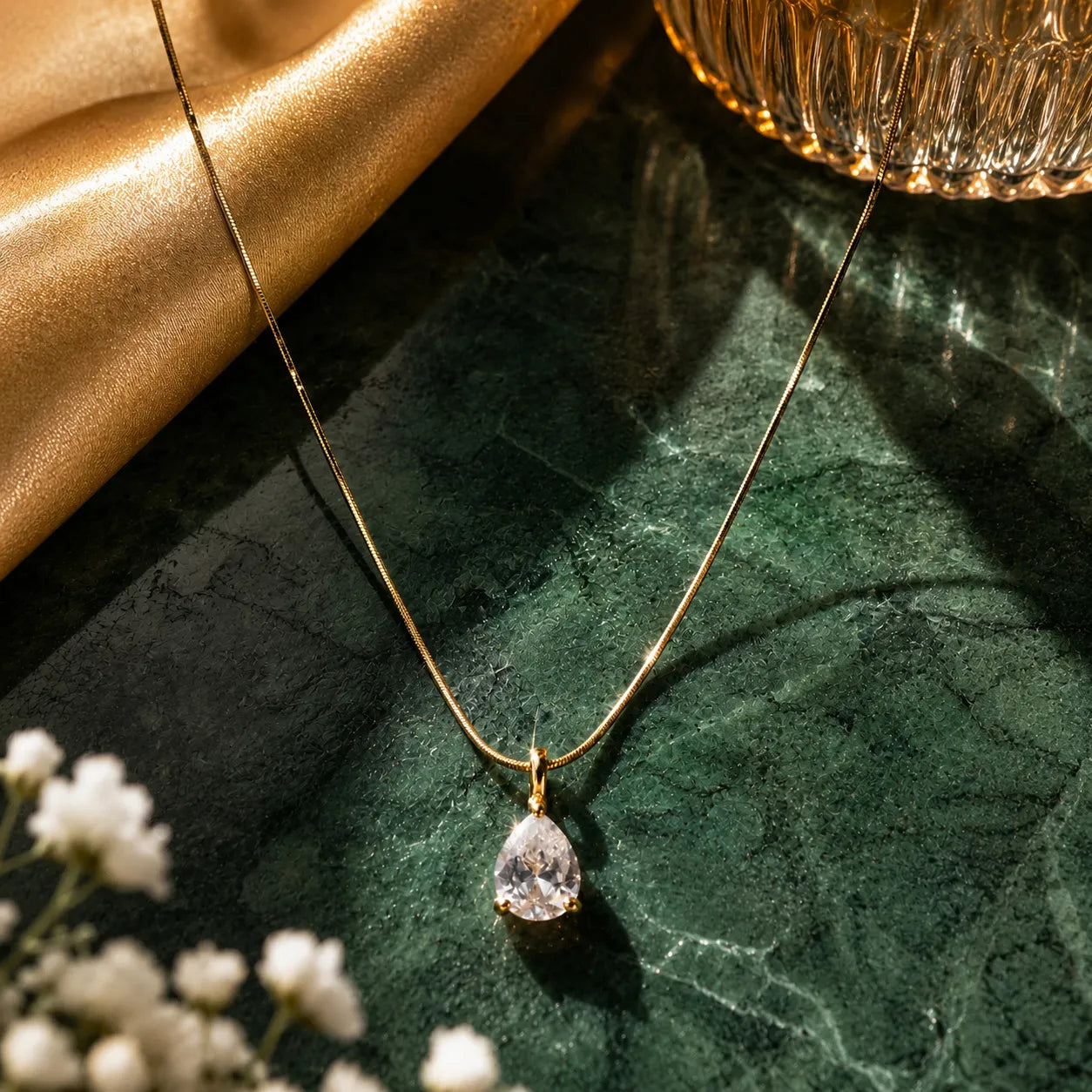 Gold necklace with a teardrop-shaped pendant on a green marble surface with white flowers and a gold cup.