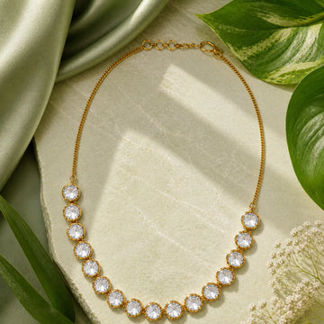 Gold necklace with clear stones on a light surface with green leaves