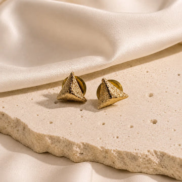 Gold pyramid-shaped earrings on a beige stone surface with soft fabric background