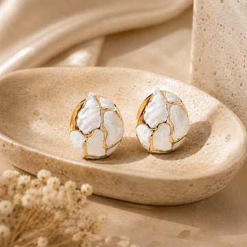 Pair of gold and white earrings on a beige stone surface with dried flowers.