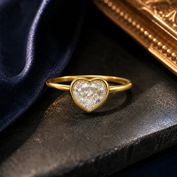 Gold heart-shaped diamond ring on a dark surface with a gold box in the background