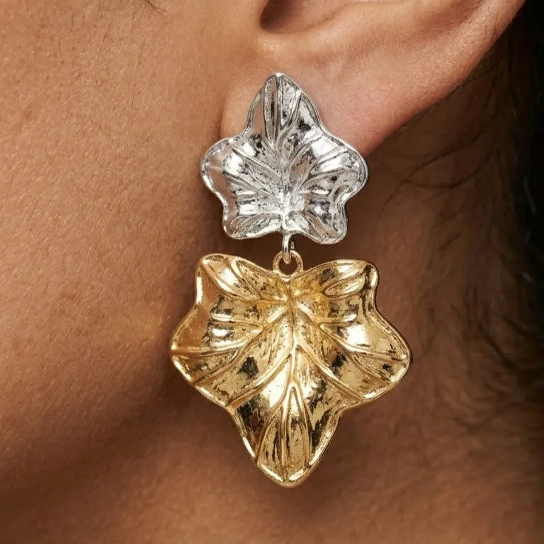 Gold and silver leaf-shaped earrings on an ear against a brown background