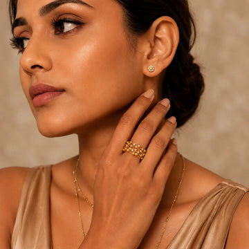 Woman wearing gold jewelry including earrings, ring, and necklace against a neutral background