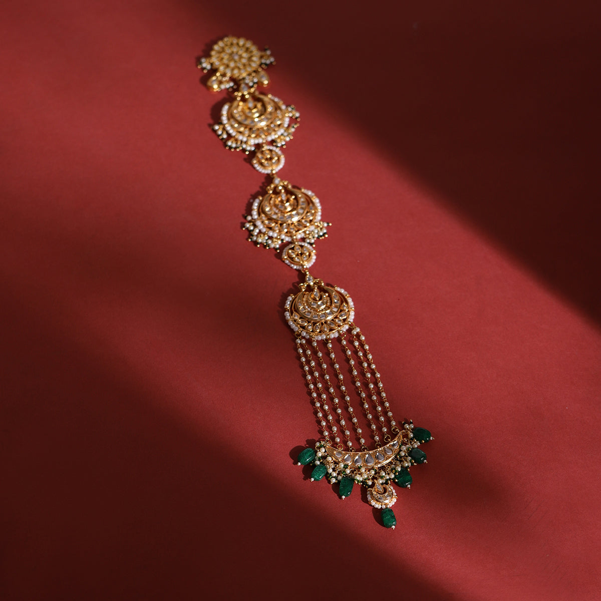 A gold-colored kundan beaded braid with ornate floral patterns and green stone accents, displayed against a red background.