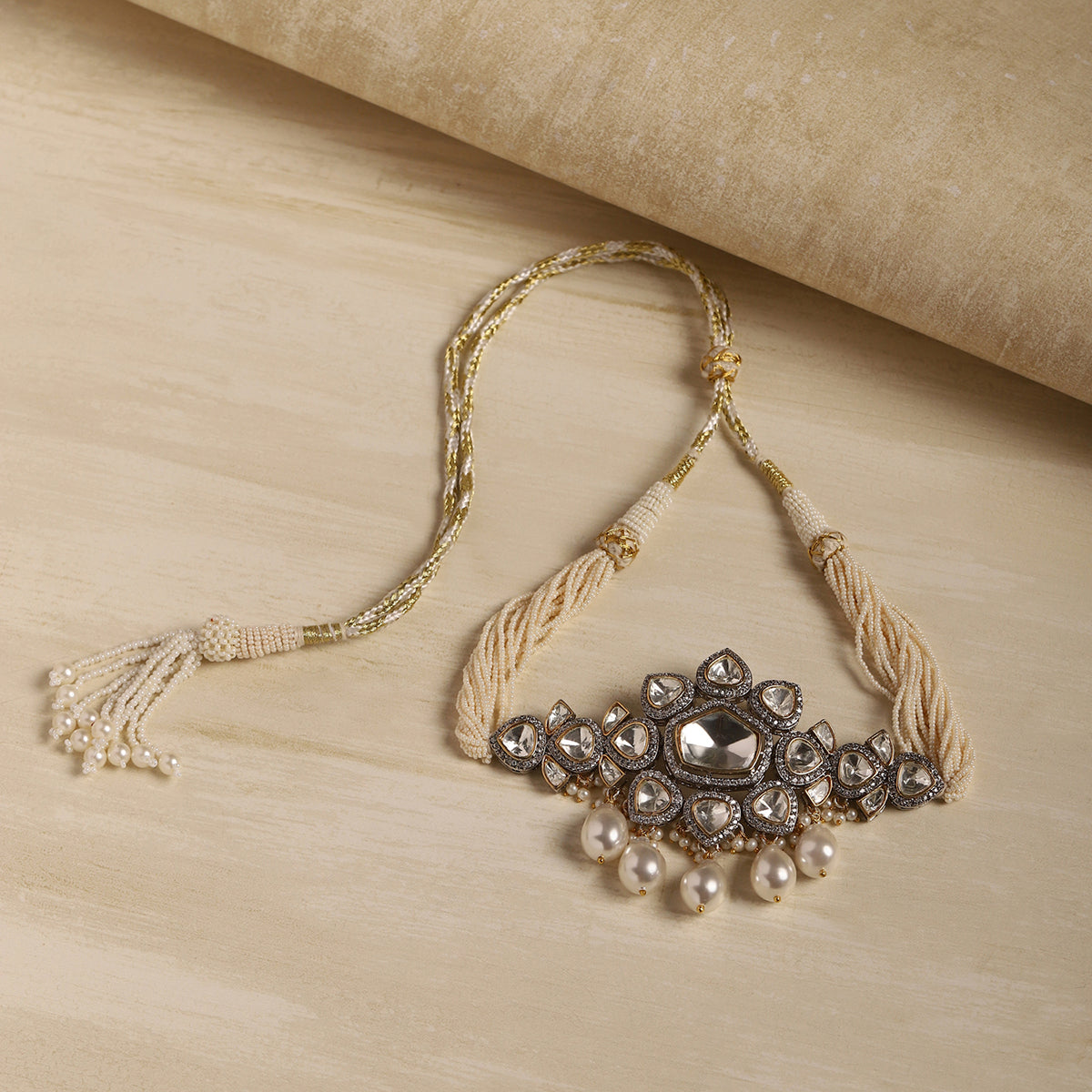 A traditional Indian silver-toned Kara necklace with a central clear stone, surrounded by small clear stones and dangling white pearls, finished with cream-colored braided cords and a tassel, displayed on a light wood surface.
