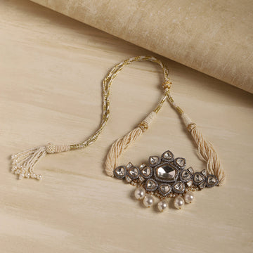A traditional Indian silver-toned Kara necklace with a central clear stone, surrounded by small clear stones and dangling white pearls, finished with cream-colored braided cords and a tassel, displayed on a light wood surface.