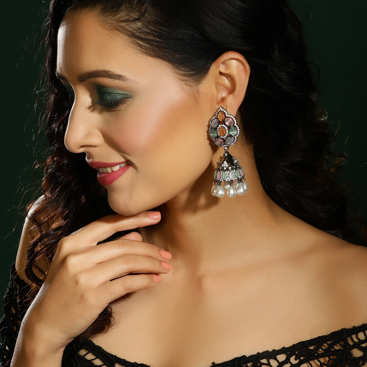 A woman wearing a large ornate silver-toned jhumka earring with multicolored stones and white pearl drops, shown in a close-up portrait against a dark green background.