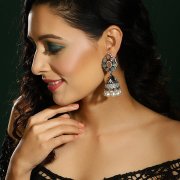 A woman wearing a large ornate silver-toned jhumka earring with multicolored stones and white pearl drops, shown in a close-up portrait against a dark green background.