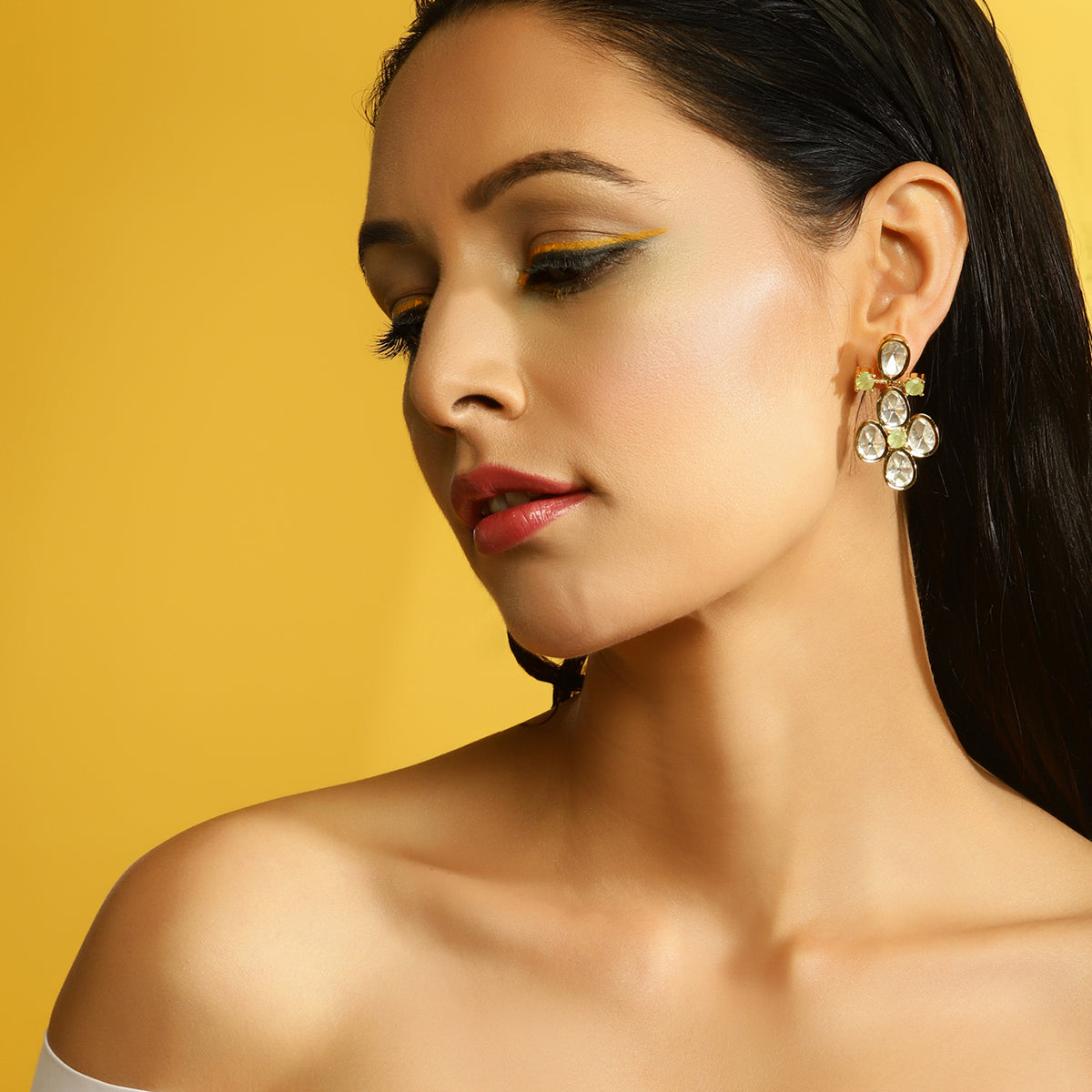 A close-up of a woman wearing a gold and crystal cluster drop earring, set against a solid yellow background.