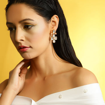 A woman with dark hair pulled back wears a dangling statement earring with clear gemstones and rose gold-toned metal, posed against a bright yellow background.