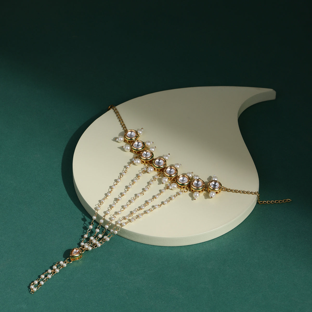 A gold-colored hand harness adorned with white pearls and precious stones, displayed on a white stand against a green background.