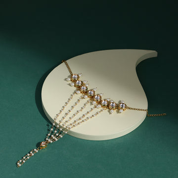 A gold-colored hand harness adorned with white pearls and precious stones, displayed on a white stand against a green background.